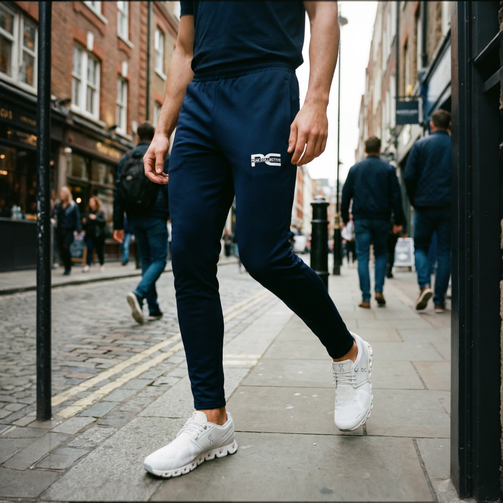 Man wearing AeroJogger men’s slim fit navy tapered jogger pants with zipped pockets and white trainers, styled for casual streetwear on a city sidewalk.
