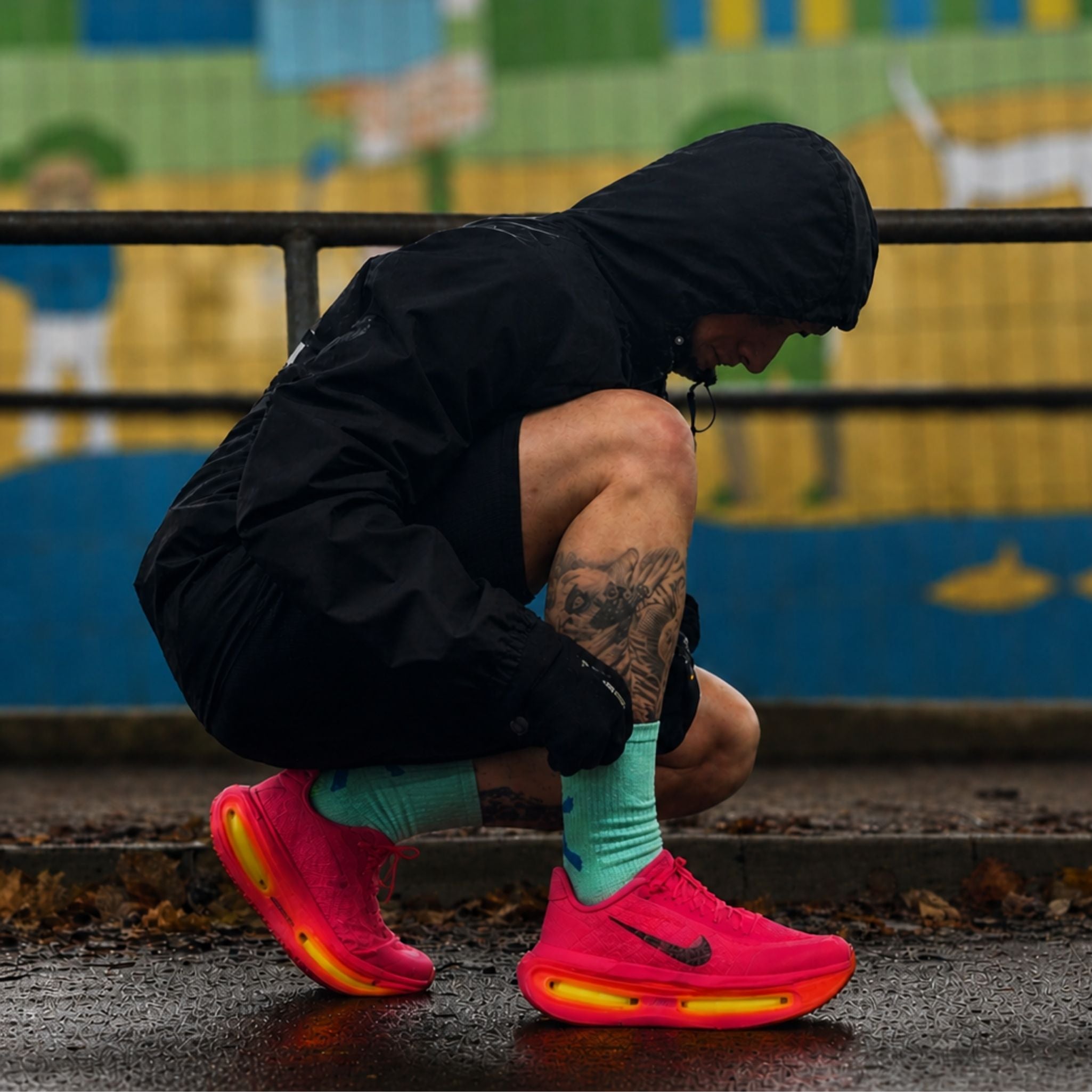 Person crouching on street wearing black AerisShell hoodie with bright pink Nike running trainers with yellow soles and Momentum green CoreSteppers – urban performance activewear outfit for running, training, and street fitness style