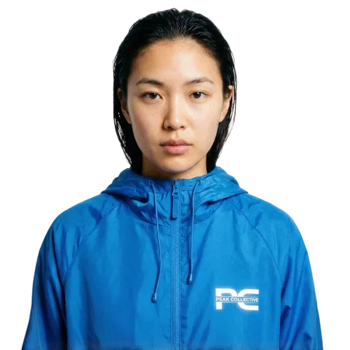 Model wearing AerisShell lightweight performance windbreaker jacket in Sapphire Blue with a visible brand logo on a grey background, designed for running, training, and outdoor activity.