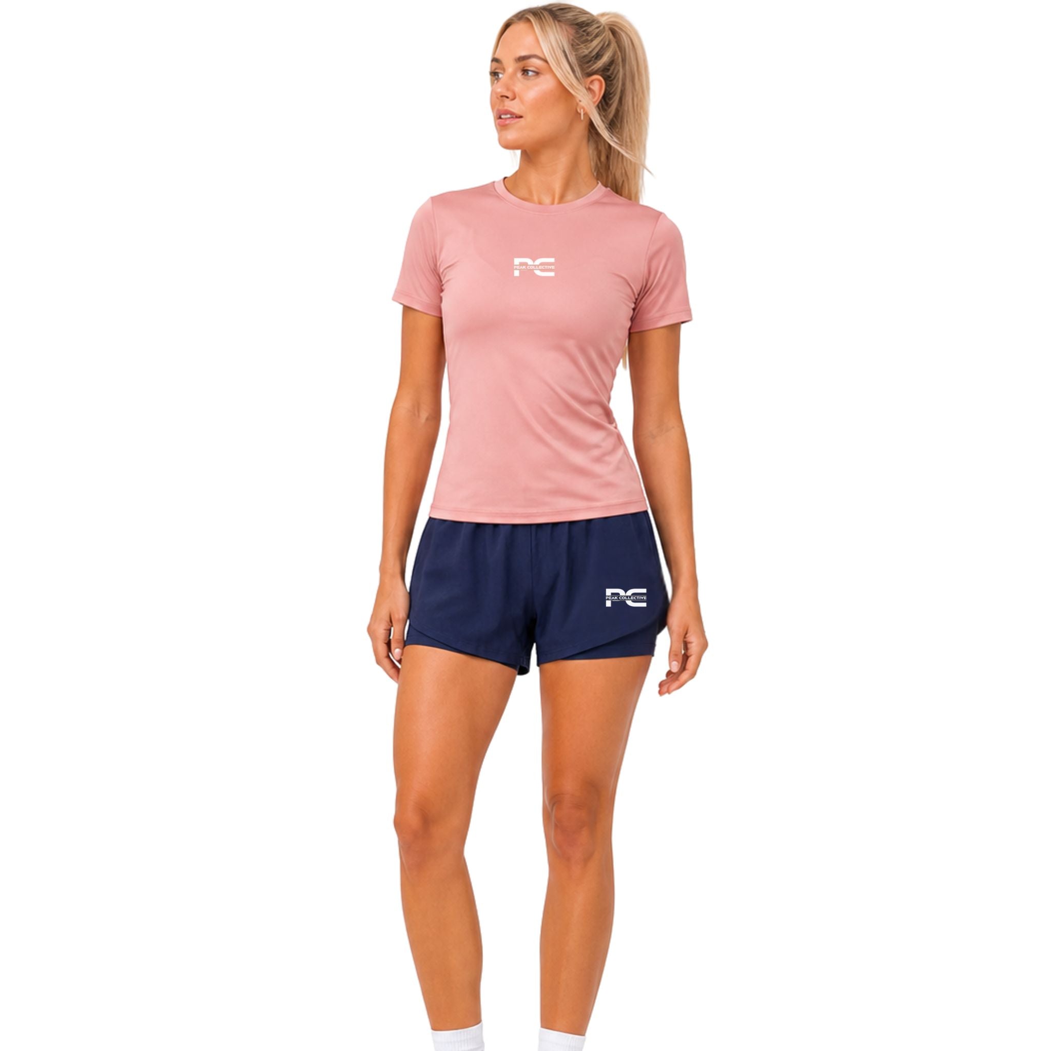 Woman wearing pink AuraTee performance t-shirt and navy LumaShorts with logo on white background – breathable women’s running outfit for gym training, jogging, fitness workouts, and everyday activewear