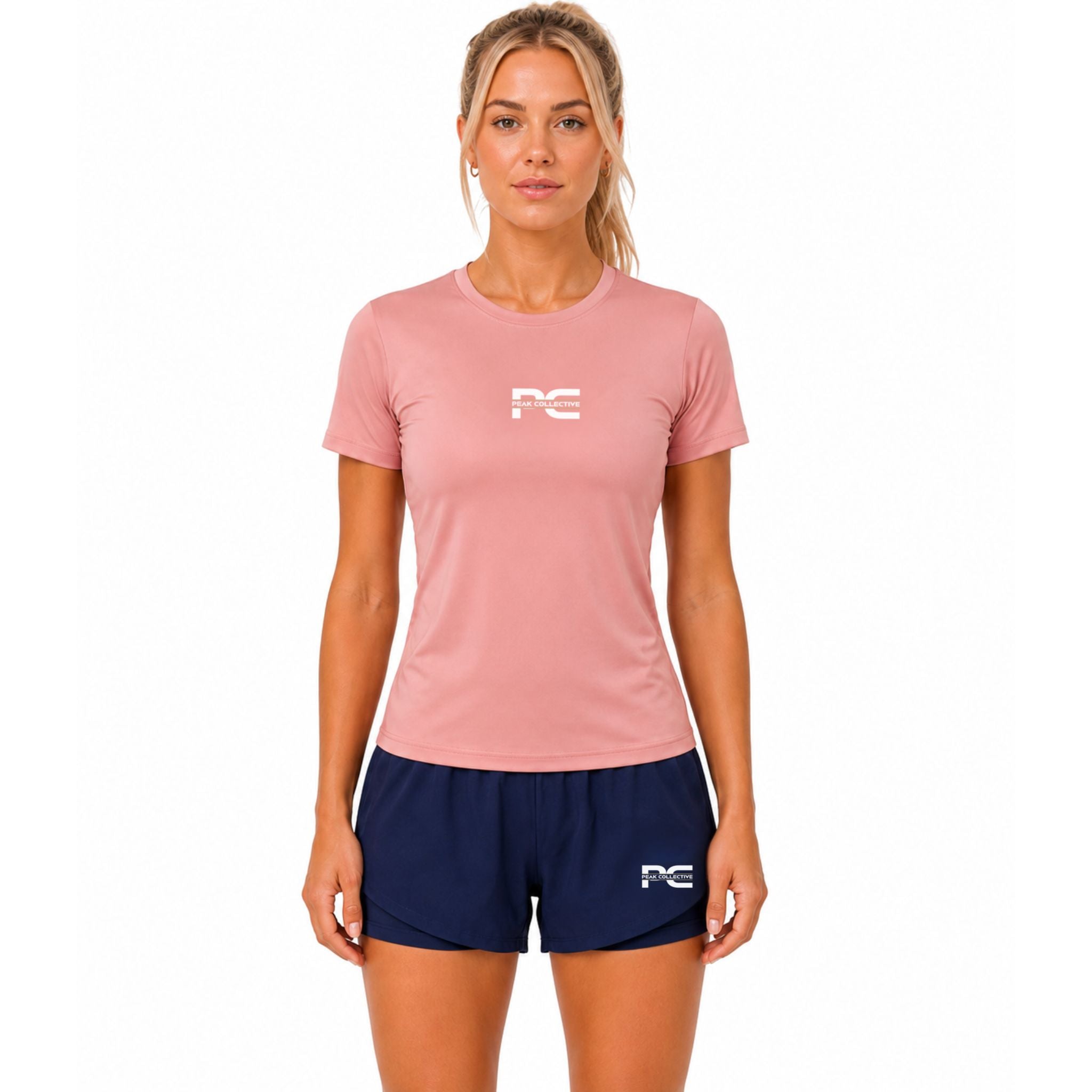 Woman wearing pink AuraTee performance t-shirt and navy LumaShorts with logo on white background – breathable women’s running outfit for gym training, jogging, fitness workouts, and everyday activewear